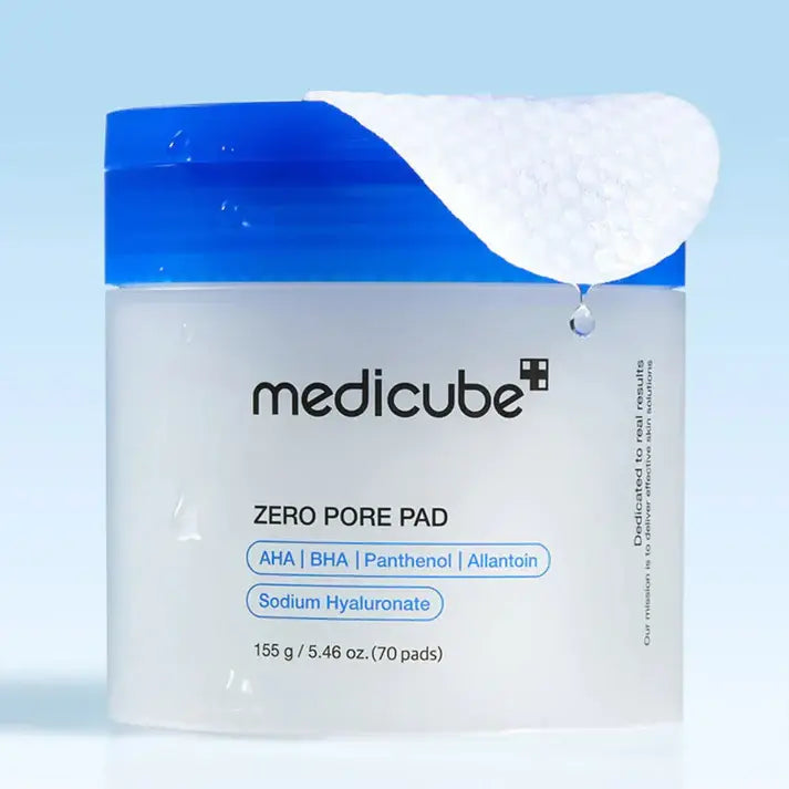 Main retirant un disque imbibé du Medicube Zero Pore Pad 2.0, avec sérum AHA/BHA s’écoulant du coton, sur fond bleu.